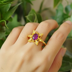 May include: A gold ring featuring a starburst design with a central oval-cut, deep purple gemstone. The ring is adorned with small, clear accent stones and intricate detailing. The ring is a piece of jewelry.