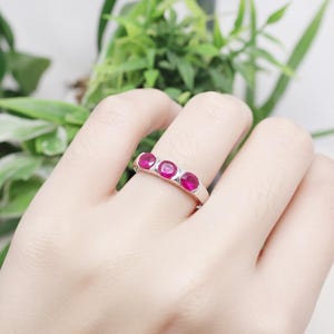 May include: A silver ring featuring three oval-cut, vibrant red gemstones. The ring is set in a simple, modern style, with the stones aligned horizontally. The gemstones are the focal point, catching the light and providing a pop of color.