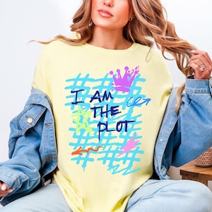 Puede incluir: Camiseta amarilla clara con el texto "I AM THE PLOT" en azul, con una corona y otros gráficos coloridos. La camiseta se combina con una chaqueta vaquera.
