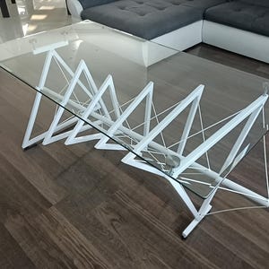 May include: A contemporary coffee table with a glass top and a distinctive white geometric base. The table has a clear glass surface supported by interconnected white triangular shapes. The table is approximately 120 cm long and 61 cm wide.