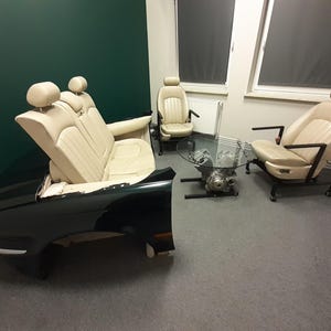 Könnte beinhalten: Ein einzigartiges Möbelset mit Autositzen und einem Sofa, das aus dem Heck eines Autos gefertigt wurde. Die Stühle und das Sofa sind mit cremefarbenem Leder bezogen. Ein Tisch mit Glasplatte steht auf einem Motorblock-Sockel.