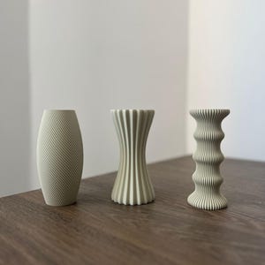 Peut inclure: Trois vases décoratifs de couleur vert clair, chacun avec un design unique, disposés sur une surface en bois brun. Le vase de gauche présente des lignes diagonales, celui du milieu une forme évasée et celui de droite un design empilé.