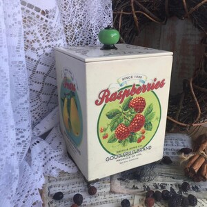 Może przedstawiać: Wytłoczona, kremowa puszka z zielonym guzikiem na pokrywie. Puszka posiada etykietę z napisem "Raspberries" w czerwonym skrypcie, zielonym okręgiem z malinami i tekstem "Since 1830".
