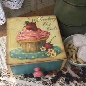 Puede incluir: Una caja de madera con un cajón, con una ilustración de cupcake con el texto "Strawberry Cup Cake". El cupcake tiene glaseado rosa y una fresa. El cajón tiene un frente azul y un pomo rosa.