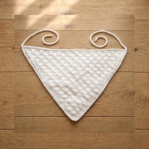 May include: A white, triangular knitted shawl with a textured pattern. The shawl has a decorative edge and two curled ties at the top. The item is displayed on a wooden surface.