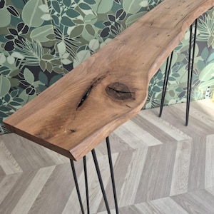 May include: A natural wood console table with a live edge and black hairpin legs. The table has a knot detail and a warm brown color. The table is approximately 60 inches long.