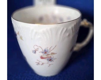 May include: A white porcelain teacup featuring a delicate floral pattern with blue and purple flowers and green leaves. The cup has a textured, embossed rim detail and a gold-toned trim along the top edge, complete with a curved handle.