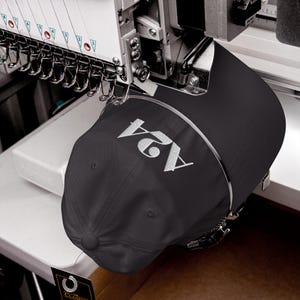 Könnte beinhalten: Eine schwarze Baseballkappe mit dem weiß gestickten Text A24 auf der Vorderseite. Die Kappe ist in eine industrielle Stickmaschine eingespannt, wobei mehrere Nadeln und Fadenführungen über der Kappenkrone sichtbar sind.
