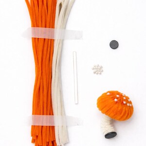 Op de afbeelding: Ambachtelijke benodigdheden voor het maken van een paddenstoelornament. Inclusief oranje en crèmekleurige pijpenragers, een klein wit stokje, een zwarte magneet, een cluster van parels en een afgewerkte oranje paddenstoel met parelaccenten.