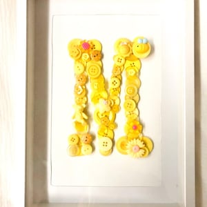 May include: A framed artwork featuring the letter "M" crafted from various yellow buttons and decorative elements. The letter is centered on a white background within a white frame. The buttons vary in size and shape, creating a textured, dimensional effect.