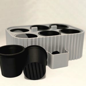May include: A gray 3D-printed planter with six circular openings, accompanied by two black plastic plant pots. The planter has a ribbed design and a small rectangular compartment. The pots are approximately 3 inches tall.