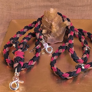 Könnte beinhalten: Geflochtene Hundeleine in Schwarz, Rot und Gold. Die Leine hat silberfarbene Metallverschlüsse und sternförmige Anhänger. Die Leine wird auf einer goldfarbenen Oberfläche präsentiert.