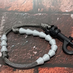 May include: A gray paracord lanyard featuring a series of smooth, conical gray beads threaded along the cord. The lanyard is finished with a black plastic swivel lobster clasp attached to one end, providing a secure point for connecting items.