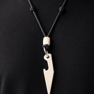 May include: A cream-colored pendant necklace with a black cord. The pendant is shaped like a knife with a serrated edge and a bottle opener. The necklace features a cylindrical bead and a metal ring.