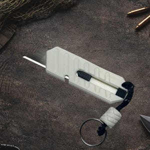 May include: A white utility knife with a black and white handle and a lanyard with a metal ring. The knife is on a dark, textured surface with other tactical gear in the background.