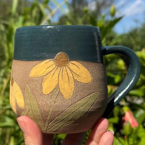 May include: A ceramic mug featuring a dark teal glazed upper section and a textured, unglazed tan lower body. The tan surface is decorated with carved and painted yellow flowers with brown centers and light green leaves on the exterior.