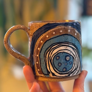 May include: Handmade ceramic mug with a brown base and handle. The mug features a unique design with concentric circles in shades of blue, brown, and white, accented with black lines and white dots. A functional and artistic piece.