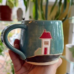 May include: A teal ceramic mug with a house design. The house has a red roof, white walls, and a red door. The mug has a large handle and a black base. The background is blurred, with green plants visible.