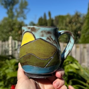 May include: Handmade ceramic mug with a landscape design. The mug features a yellow sun, green hills, and a blue lake against a turquoise sky. The handle and rim have a turquoise glaze. The mug is held in a hand.