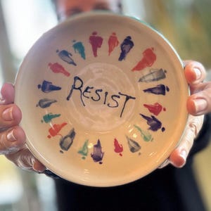 May include: A ceramic bowl with a cream-colored interior and a turquoise rim. The word "RESIST" is hand-lettered in black in the center. Around the rim are colorful, abstract figures in various colors, including red, blue, and green.