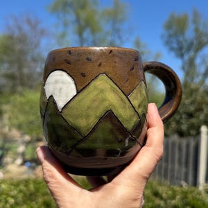 May include: A handmade ceramic mug with a mountain landscape design. The mug features a brown top with dark brown accents, green mountains, and a white moon. The handle and base are brown.