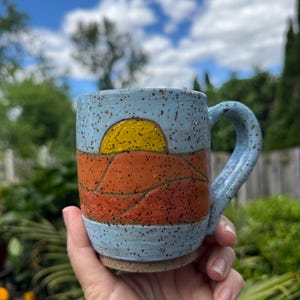 May include: A ceramic mug with a speckled light blue glaze featuring a hand-painted landscape design. The design includes a bright yellow sun rising over layered orange hills. The mug has a sturdy handle and a natural, unglazed brown clay base.
