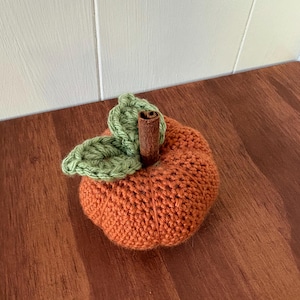 May include: A crocheted orange pumpkin with a textured, ribbed surface. The top features two light green crocheted leaves and a central brown cinnamon stick stem, creating a rounded, soft-sculpted appearance with visible yarn stitches.