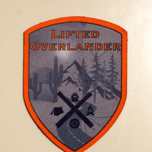 Könnte beinhalten: Ein schildförmiger Aufnäher mit einem orangefarbenen Rand und den Worten "LIFTED OVERLANDER" in Orange. Der Innenraum zeigt einen grauen Hintergrund mit Silhouetten von Bergen, Bäumen, einer Straße, einem Zelt, einem Lagerfeuer und einem Pfotenabdruck.