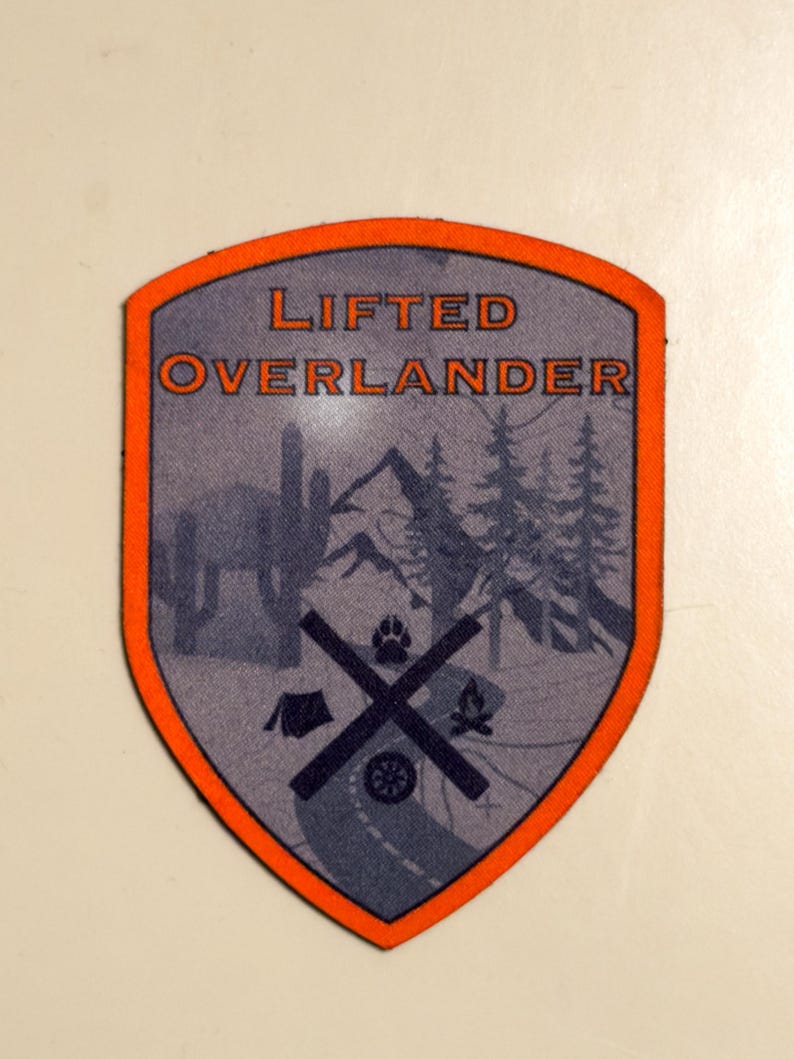 May include: A shield-shaped patch with an orange border and the words "LIFTED OVERLANDER" in orange. The interior features a gray background with silhouettes of mountains, trees, a road, a tent, a campfire, and a paw print.