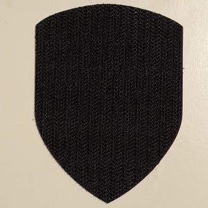 May include: A black shield-shaped patch with a textured surface. The patch has a rounded top and a pointed bottom. The background is a light beige color.