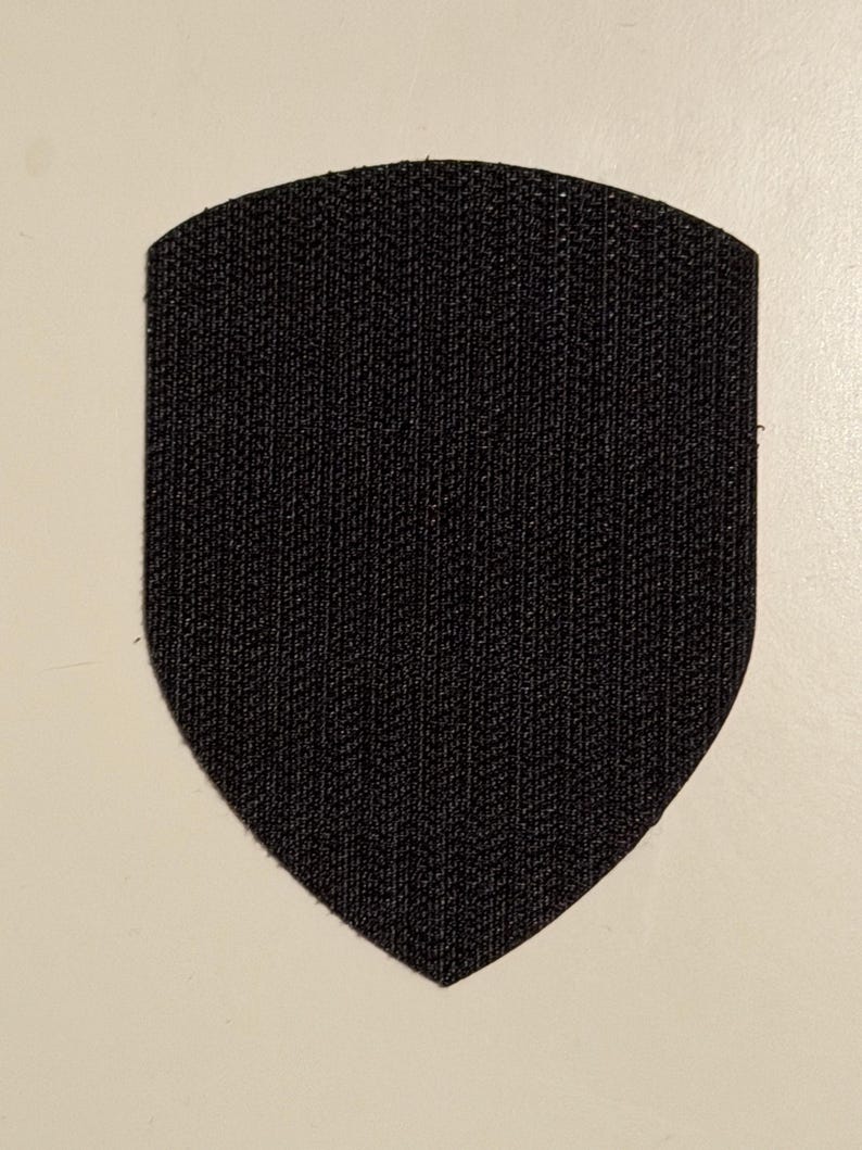 May include: A black shield-shaped patch with a textured surface. The patch has a rounded top and a pointed bottom. The background is a light beige color.