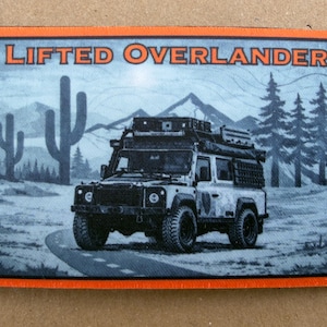Könnte beinhalten: Rechteckiger Aufnäher mit orangefarbenem Rand. Die Schwarz-Weiß-Illustration zeigt ein geländegängiges Fahrzeug auf einer Straße, mit dem Text "LIFTED OVERLANDER" darüber. Der Hintergrund zeigt Berge, Kakteen und Kiefern.