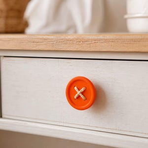 Op de afbeelding: Close-up van een wit geschilderde lade met een oranje knopvormige knop. De knop heeft een kruissteekontwerp van natuurlijk gekleurd touw. De lade maakt deel uit van een lichtgekleurde houten bureau of dressoir.