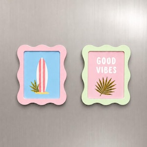 Può includere: Due magneti rettangolari con bordi ondulati. Il magnete a sinistra presenta una tavola da surf rosa e bianca su sfondo azzurro. Il magnete a destra ha uno sfondo rosa chiaro con il testo bianco GOOD VIBES sopra un'illustrazione di una foglia di palma verde.
