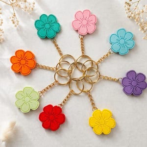 Peut inclure: Un ensemble de huit porte-clés en forme de fleurs de différentes couleurs : turquoise, rose, bleu, orange, vert, rouge, jaune et violet. Chaque fleur est attachée à une chaîne et un porte-clés dorés. Les porte-clés sont disposés en cercle.