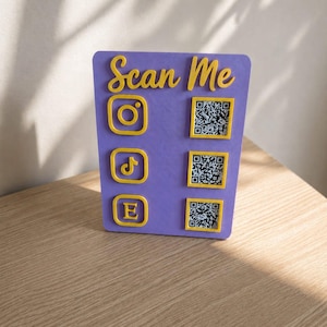 Peut inclure: Panneau violet avec l'inscription "Scan Me" en jaune. Il présente des icônes jaunes pour Instagram, TikTok et Etsy, ainsi que des codes QR correspondants.