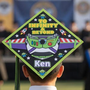 Puede incluir: Un birrete de graduación decorado con un diseño de Buzz Lightyear, con la frase "To Infinity & Beyond". El birrete es negro con un diseño verde, negro y blanco. El nombre "Ken" está impreso en el birrete. Una borla cuelga del birrete.