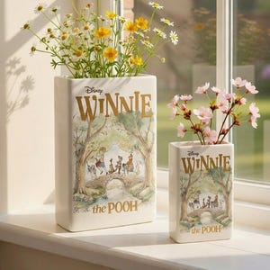 Könnte beinhalten: Zwei rechteckige Winnie-Puuh-Vasen mit Blumen. Die größere Vase hat gelbe und weiße Blumen, und die kleinere Vase hat rosa Blumen. Beide Vasen tragen die Worte "Disney Winnie the Pooh" in goldfarbenen Buchstaben und eine Szene von Puuh und seinen Freunden.