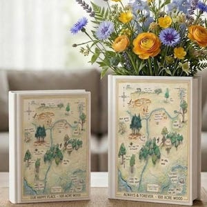 May include: Two white book-shaped vases with a map design, one holding a vibrant floral arrangement. The map features trees, a river, and text that reads "OUR HAPPY PLACE - 100 ACRE WOOD" and "ALWAYS & FOREVER - 100 ACRE WOOD".