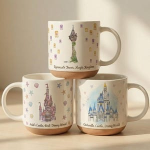 Op de afbeelding: Drie witte keramische mokken met bruine bodems. Elke mok heeft een ander Disney-kasteelontwerp: Rapunzels Toren, Ariëls Kasteel en Cinderella Castle. De mokken hebben gebogen handvatten en zijn versierd met pastelkleuren en de tekst "Rapunzel's Tower, Magic Kingdom", "Ariel's Castle, Walt Disney World" en "Cinderella Castle, Disney World".