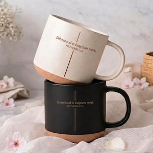 May include: Two ceramic mugs, one cream-colored and one black, stacked together. The cream mug has a brown base and the text "fatherhood is kingdom work PROVERBS 22:6". The black mug has the same text. Both mugs have a handle.