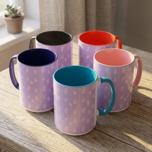May include: A set of four ceramic mugs with a light purple exterior featuring a repeating white leaf pattern. Each mug has a distinct interior and handle color, including dark blue, black, orange, and light pink, with one teal-handled mug.