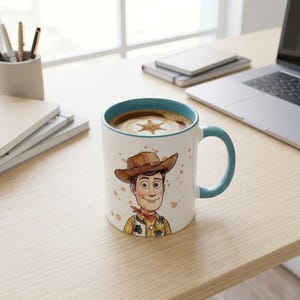 May include: A white ceramic mug with a teal interior and handle, featuring a colorful illustration of a cowboy character wearing a brown hat, yellow shirt, and cow-print vest, with a star-shaped foam pattern visible in the coffee inside.