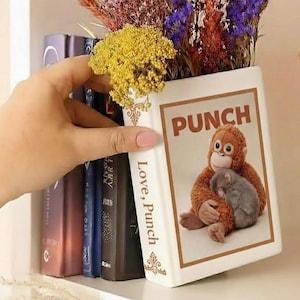 May include: A white ceramic vase shaped like a book, with the word "PUNCH" and an illustration of an orangutan hugging a mouse. The vase is filled with colorful dried flowers. The spine of the book reads "Love, Punch".