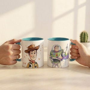 May include: Two white ceramic mugs with teal interiors and handles. One mug features a colorful illustration of a cowboy character wearing a brown hat and yellow shirt. The other mug displays a colorful illustration of a space ranger character.