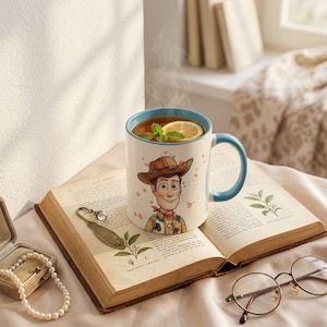 May include: A white ceramic mug with a light blue interior and handle, featuring a cartoon illustration of a cowboy character wearing a brown hat, yellow shirt, and red neckerchief, filled with steaming tea, a lemon slice, and mint leaves.