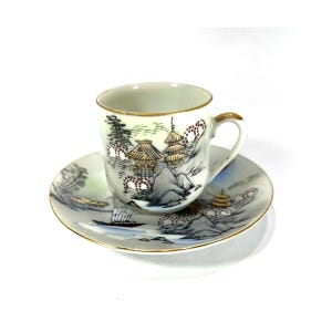 May include: A small, white porcelain teacup and saucer set. The cup and saucer are decorated with a hand-painted landscape scene featuring mountains, pagodas, and a boat. Gold trim accents the edges of both pieces, adding a touch of elegance.