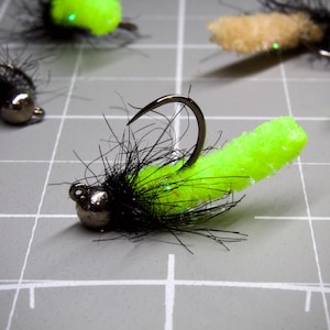 May include: Close-up of several fishing flies. Each fly features a silver bead head, a black body with wispy fibers, and a bright neon green or tan foam body. The flies are arranged on a gray grid surface.