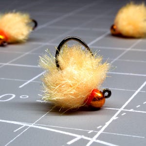 May include: Close-up of three fishing flies. Each fly features a fluffy, light-yellow body, a small, orange bead, and a black hook. The flies are arranged on a gray surface with white grid lines, suggesting a crafting or work area.