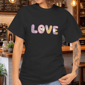 May include: A black short-sleeved t-shirt featuring the word LOVE printed in a pastel-colored, patchwork-style font. Each letter is composed of different soft-toned patterns, including light pink, yellow, and lavender, with a small heart icon.